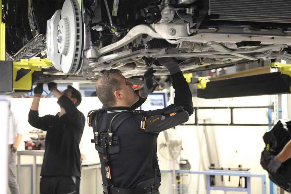 Assembly line workers reaching overhead under a vehicle, one wearing a shoulder exoskeleton