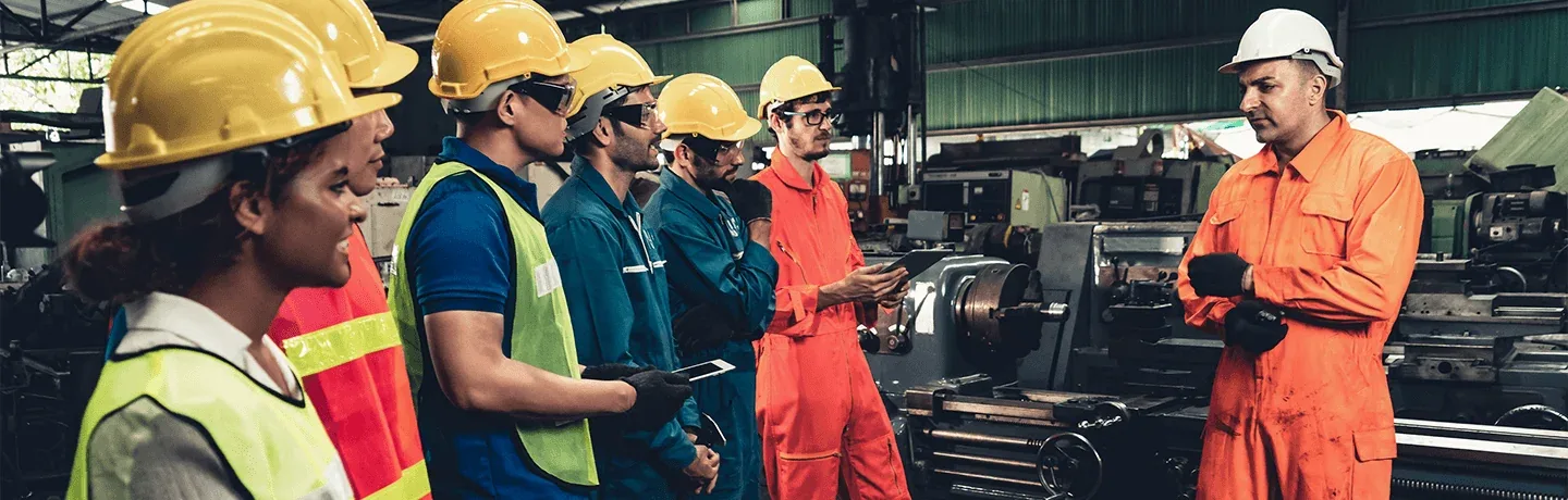 Employees in hard hats and hi-vis clothing receiving a safety briefing on a factory floor