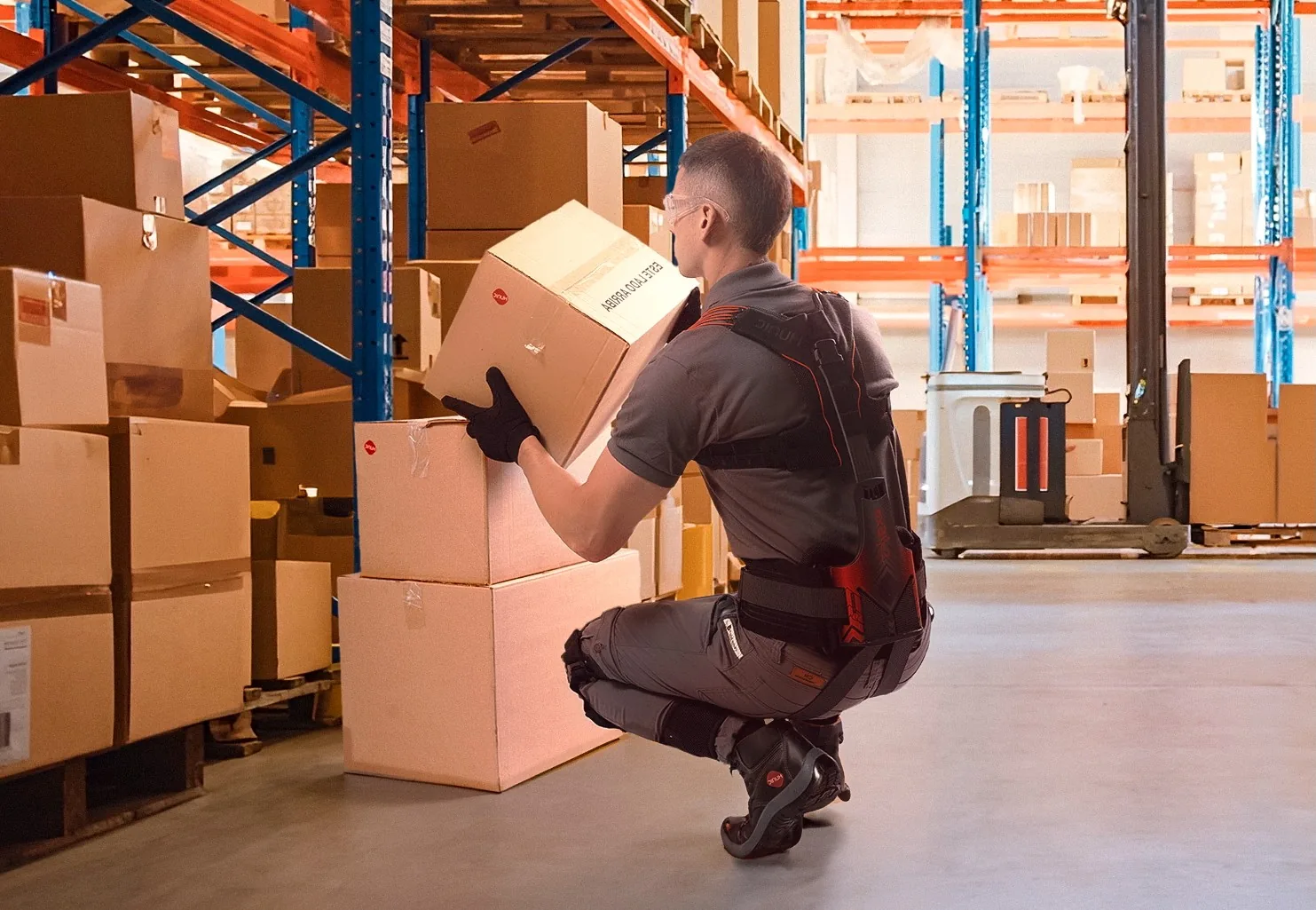 Warehouse worker crouching to lift a box from a low shelf while wearing a back-support exoskeleton